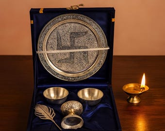 Puede incluir: Un juego religioso plateado en una caja de terciopelo azul. El juego incluye un plato decorativo con un diseño de esvástica, dos cuencos pequeños, una pluma decorativa y una copa pequeña. A la derecha de la caja hay una lámpara de aceite de latón encendida.