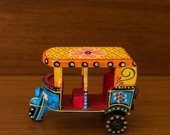 May include: A colorful, miniature wooden auto-rickshaw. The vehicle features a yellow roof with a floral design, blue and red body, and black wheels. The decorative piece is painted with intricate patterns and details.