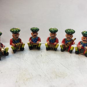 Könnte beinhalten: Eine Sammlung von sechs kleinen, handbemalten Holzfiguren von Musikern. Jede Figur trägt einen grün-gelben Turban, eine rote Jacke, ein blaues Hemd und eine gelbe Hose. Die Musiker spielen verschiedene Instrumente, darunter Trommeln und Saiteninstrumente.