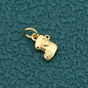 May include: A small, gold-colored charm in the shape of a mitten. The charm has a textured surface and a small loop at the top for attaching to a chain or bracelet. The mitten has a snowflake design on the front.