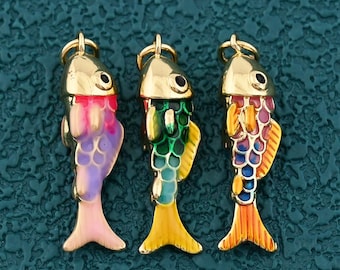 May include: Three colorful fish charms with gold-colored caps and eye details. The fish are painted in shades of pink, purple, green, yellow, and orange. Each charm has a small loop at the top for attaching to a necklace or bracelet.