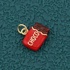 May include: A red and brown chocolate bar charm with the word "CHOCO" in white lettering. The charm has a gold-colored loop for attaching to a necklace or bracelet. The background is a textured teal surface.