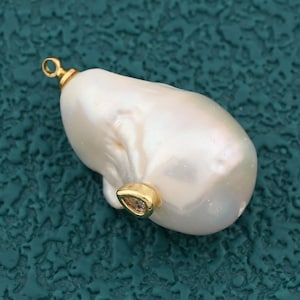 May include: A large, irregular-shaped white pearl pendant with a gold-colored bail. The pearl has a smooth, lustrous surface and a slightly off-white hue. The pendant is designed for jewelry making.