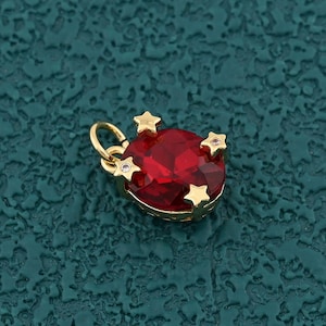 Op de afbeelding: Een goudkleurige hanger met een grote, ovale, rode edelsteen in het midden. De hanger is versierd met kleine, goudkleurige steraccenten en een lus voor een ketting. De achtergrond is een getextureerde teal kleur.