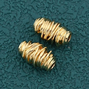 Puede incluir: Dos cuentas doradas en forma de barril con un diseño de alambre texturizado y enrollado. Las cuentas están colocadas sobre una superficie texturizada de color verde azulado. Estas cuentas son adecuadas para la fabricación de joyas.