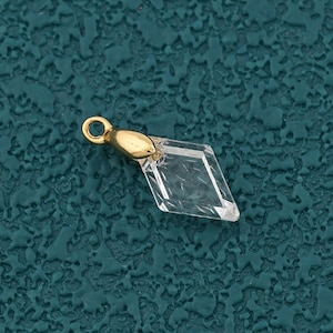 May include: A clear, diamond-shaped crystal charm with a gold-colored metal loop and cap. The charm is set against a textured teal background. This small pendant is suitable for jewelry making or crafting.