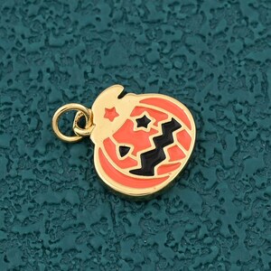 Puede incluir: Un dije de calabaza de color dorado con esmalte naranja y rasgos faciales negros. El dije tiene un pequeño lazo para sujetarlo a un collar o pulsera. El diseño de la calabaza incluye recortes en forma de estrella.