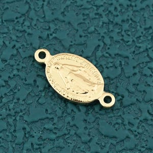 May include: A gold-colored oval charm with a religious image, featuring two small loops for attachment. The charm is set against a textured teal background. The image depicts a religious figure.