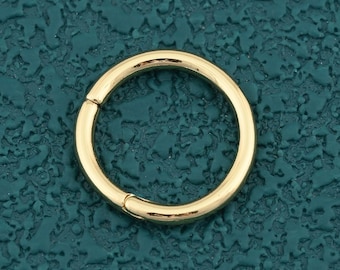 Pode incluir: Um piercing de nariz ou brinco articulado dourado. A joia circular é mostrada contra um fundo texturizado azul-petróleo. O anel tem um design simples e sem costuras.