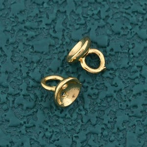 May include: Two small, gold-colored jewelry findings with a cap and loop design. The findings are on a textured teal background. The caps are rounded and have a small loop attached for stringing or connecting to other jewelry components.