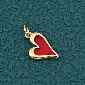May include: A gold-colored heart-shaped charm with a red enamel filling. The charm has a small loop at the top for attaching to a necklace or bracelet. The charm is on a textured teal background.
