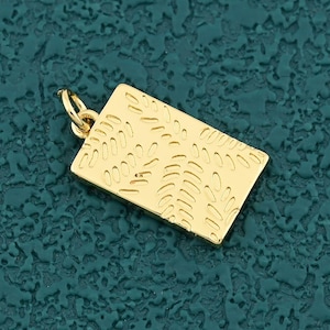 May include: A gold-colored rectangular pendant with a textured surface and a small loop for a chain. The pendant has a decorative pattern of curved lines and is set against a teal background.