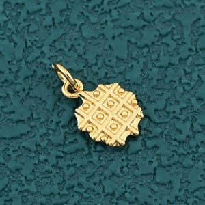 May include: A small, gold-colored charm with a waffle design. The charm has a textured surface with a grid pattern of squares and circles. It has a small loop at the top for attaching to a necklace or bracelet. The charm is set against a teal background.