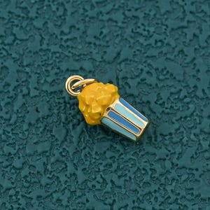 May include: A gold-colored popcorn charm with yellow popcorn and a blue and white striped container. The charm has a small loop at the top for attaching to a necklace or bracelet. The charm is small and detailed.