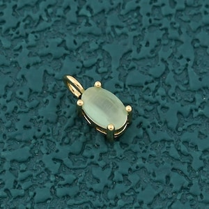 May include: A gold pendant with a light green oval-shaped gemstone. The gemstone is held in place by four gold prongs. The pendant has a small loop at the top for a chain. The background is a textured teal color.