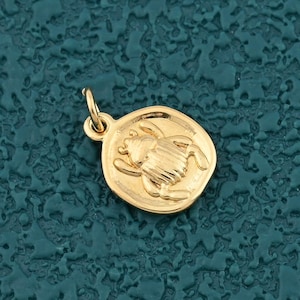 May include: A small, gold-colored pendant with a circular design. The pendant features a detailed relief of a scarab beetle. The beetle is centered within the circle, and the pendant has a small loop for attaching to a chain.