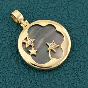 May include: Gold-tone pendant featuring a celestial design. The round pendant has a dark, iridescent center with a crescent moon shape and three star cutouts. Two stars are embellished with small, clear gemstones.