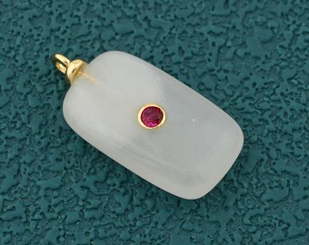 Puede incluir: Un colgante rectangular con una fianza dorada y una pequeña gema roja redonda en el centro. El colgante es de un color claro y translúcido, posiblemente blanco o blanquecino.