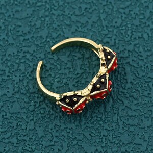 Könnte beinhalten: Ein goldfarbener Ring mit einem einzigartigen Design. Der Ring hat drei dekorative Elemente mit einem schwarz-roten Muster. Der Ring ist offen, was eine Größenanpassung ermöglicht. Der Ring ist vor einem strukturierten tealfarbenen Hintergrund platziert.