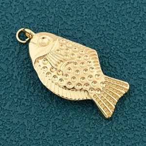 Puede incluir: Un colgante de pez dorado con una superficie texturizada. El pez tiene un diseño detallado, que incluye escamas y aletas, y un pequeño lazo en la parte superior para sujetarlo a una cadena o cordón. El colgante está sobre un fondo verde azulado.