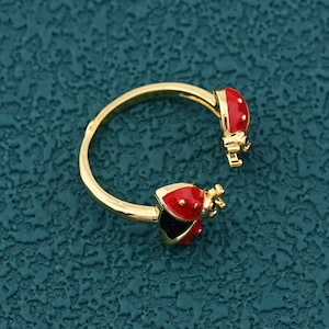 May include: A gold-colored adjustable ring featuring two red ladybug designs with black spots and accents. The ring is open-ended, allowing for size adjustment. The ladybugs have black undersides.