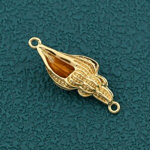 May include: A gold-colored seashell charm with a brown center. The charm has detailed ridges and two small loops for attaching to jewelry. The seashell is set against a textured teal background.