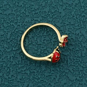 May include: A gold ring with a ladybug design. The ring features a red and black ladybug charm and a red flower. The ring is set against a textured teal background.