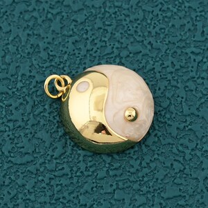 May include: A gold-plated pendant with a yin-yang symbol. The circular pendant has a polished gold finish and a white and cream-coloured swirl pattern. It includes a small loop for attaching to a chain or bracelet.