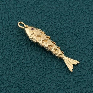 May include: A gold-colored fish pendant with a textured body and a small red gem for an eye. The fish has a loop at the top for hanging and a detailed tail fin. The pendant is set against a dark teal background.