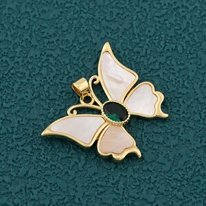 May include: A gold-colored butterfly pendant with white and light pink wings, and a green gemstone center. The pendant has a small loop for a chain. The background is a textured teal color.