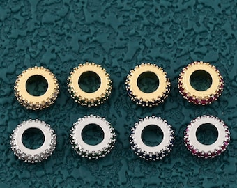 May include: Eight small, round jewelry beads in gold and silver tones, with a central hole. Some beads have red or dark blue accents. The beads are arranged in two rows of four against a textured, dark teal background.