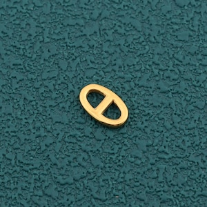 May include: A small, gold-colored metal charm shaped like an oval link with a horizontal bar across the center. The piece features a smooth, polished finish and a distinct, symmetrical design, presenting a clean and simple geometric form.