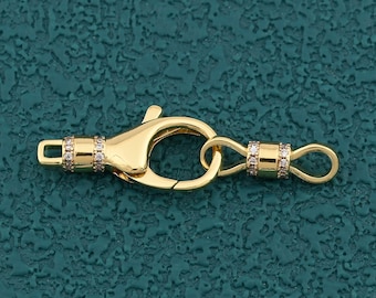 Pode incluir: Um fecho de joia dourado com um fecho de lagosta e um pequeno laço na extremidade oposta. O fecho e o laço são adornados com pequenas pedras preciosas transparentes. O fundo é de cor verde-azulado texturizado.