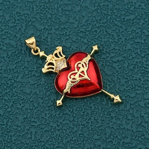 Pode incluir: Pingente dourado com um coração vermelho perfurado por duas flechas. O coração tem um design dourado e é encimado por uma coroa. O pingente está sobre uma superfície texturizada azul-petróleo.