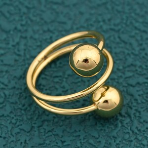 May include: A gold-colored ring with a spiral design. The ring features two spherical accents, one at the top and one at the bottom of the spiral. The ring is set against a teal-colored background.