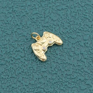 May include: A gold-toned metal charm shaped like a video game controller, featuring a small jump ring at the top for attachment. The surface of the charm is detailed with embossed button patterns and two small, clear, round inset stones.