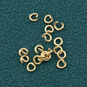 May include: A collection of small, gold-colored C-shaped jewelry findings and a few small gold chain links are scattered on a textured teal surface. The findings are open at one end, ready to connect to other jewelry components.