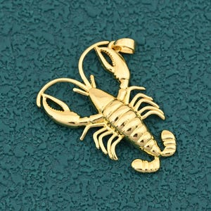 Könnte beinhalten: Ein goldfarbener Hummer-Anhänger mit detaillierten Scheren, Beinen und Körpersegmenten. Der Anhänger hat oben eine Schlaufe für eine Kette. Der Hummer ist vor einem strukturierten, teal-farbenen Hintergrund platziert.