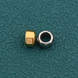 May include: Two small, cylindrical beads with a smooth, metallic finish. One bead is gold-toned and features a square-shaped profile, while the other is silver-toned and has a hexagonal-shaped outer edge with a circular hole in the centre.
