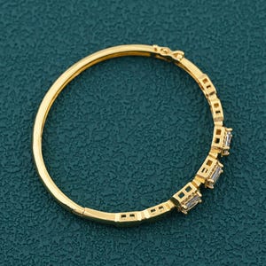May include: A gold-coloured bangle bracelet with a hinged clasp. The bracelet features a series of rectangular, clear gemstones set in a decorative pattern. The bracelet is shown against a teal-coloured background.