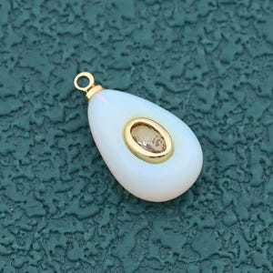 May include: A teardrop-shaped pendant with a white, opaque body and a gold-colored bail. The center features a small, oval-shaped, light-brown stone set within a gold-colored frame. The pendant is designed for jewelry making.