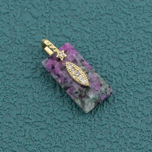 Puede incluir: Un colgante rectangular con una base de piedra moteada en tonos morados y grises. La parte superior del colgante está adornada con una anilla de metal dorado en forma de pequeña estrella, unida a un dije dorado en forma de marquesa con cristales claros.