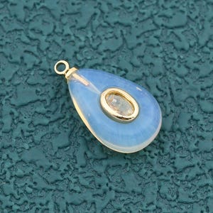May include: A teardrop-shaped pendant with a light blue, opalescent stone and a gold-colored metal setting. The center features a smaller, oval-shaped stone. A small gold loop is at the top for attaching to a chain or cord.