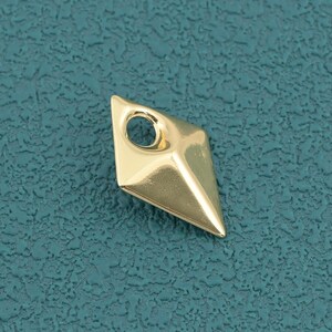 May include: A small, gold-colored metal charm shaped like a diamond or kite with a smooth, reflective surface. The charm features a single circular hole at the top for attachment, with a pointed bottom edge and faceted sides that create a 3D effect.