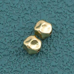 May include: Two small, metallic gold-colored beads with a smooth, slightly irregular cube-like shape. Each bead features rounded edges and a reflective surface that catches the light, creating bright highlights across their polished finish.