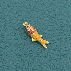May include: A small gold-toned metal charm shaped like a koi fish, featuring vibrant enamel detailing in shades of orange, yellow, and red across its scales and fins, with a small loop at the top for attachment to jewelry or other accessories.