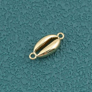 Pode incluir: Um pendente de metal em tom dourado com formato de concha de búzios, apresentando uma superfície lisa e polida com uma fenda central. O pendente inclui um pequeno elo circular em cada extremidade para fixação em joias.