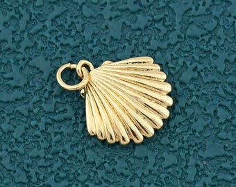 Puede incluir: Un pequeño dije de concha dorada con un lazo en la parte superior para sujetarlo a un collar o pulsera. El dije tiene un diseño detallado y acanalado y está sobre un fondo texturizado color turquesa.