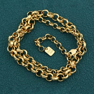 May include: A gold-colored chain necklace with a textured, rounded link design. The necklace features a small, square charm that reads "Good Luck". The chain is displayed on a teal-colored surface.