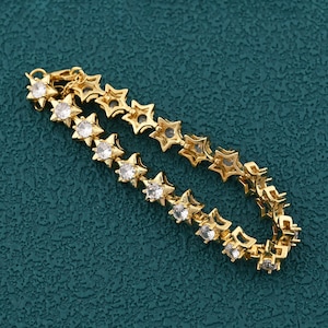 Puede incluir: Una pulsera dorada con un diseño de estrellas repetidas. Cada estrella está engastada con una piedra transparente y brillante. La pulsera se muestra sobre una superficie de color turquesa, resaltando sus detalles.